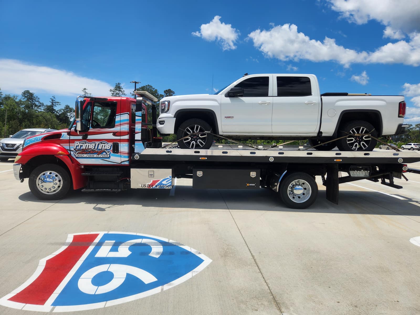 Red Prime Time Towing flatbed hauling a white GMC pickup under a blue sky, US DOT 4569907 visible on the truck